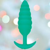 The B-Vibe Texture Vibrating Butt Plug Twist - Green (Large), a ribbed, body-safe silicone plug with a flared base, is centered against a softly blurred pastel lights background.
